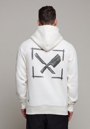 Mand iført hvid hættetrøje med sort krydset barberkniv og slagteknivsgrafik på ryggen, stående mod en lysegrå baggrund.