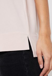 Ljusrosa topp med en mjuk textur, med sidofickor vid fållen och en avslappnad passform, tillsammans med mörka byxor.