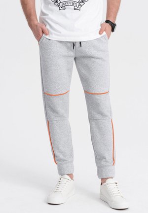 Graue Sweatpants aus weichem Stoff, mit orangefarbenen Akzenten an Taschen und Nähten sowie engeren Bündchen. Getragen mit weißen Sneakern.