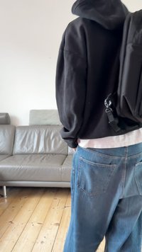 Crni ruksak s podesivim naramenicama, položen preko tamne huppere i bijele košulje. Svijetloplave traperice s vidljivim stražnjim džepovima.