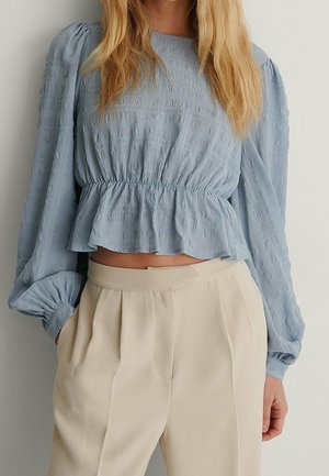 Kobieta w jasnoniebieskiej, teksturowanej bluzce z długim rękawem i marszczonym pasem oraz beżowych, plisowanych spodniach z wysokim stanem, stoi z jedną ręką w kieszeni.