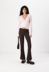 Leicht rosa Spitzenwickeloberteil mit langen Ärmeln, kombiniert mit braunen Schlaghosen mit Knöpfen und schwarzen Plateauschuhen. Schwarze Handtasche.