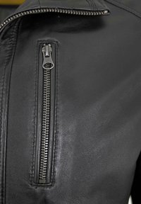 Schwarze Lederjacke mit einer diagonal verlaufenden Reißverschlusstasche, ausgestattet mit silbernen Beschlägen und einer strukturierten Oberfläche, die handwerkliches Können und Detailgenauigkeit hervorhebt.
