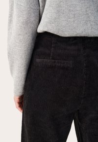Pantalon en velours côtelé noir avec texture côtelée verticale, doté d'une poche arrière, porté avec un pull gris clair.