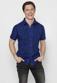 Camicia blu a maniche corte con motivo astratto blu scuro, bottoni frontali e colletto a punta, abbinata a jeans scuri.