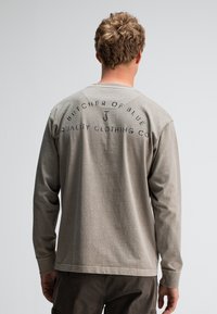 Langärmliges, hellgraues Hemd aus weichem Material. Es hat bedruckten Text auf der Rückseite mit dunklen Akzenten und einem Rundhalsausschnitt.