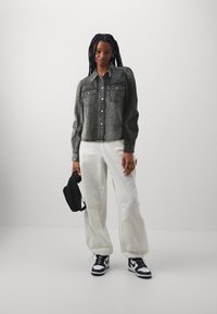 Denim jack in gewassen donkergrijs met een knoopsluiting aan de voorkant, twee borstzakken en geplooide mouwen, gecombineerd met losse witte broek en zwarte sneakers.