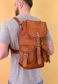 Mochila de cuero marrón con correas ajustables, solapa frontal y dos bolsillos exteriores, con herrajes plateados y acabado texturizado.