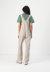 Beige overall met brede benen, verstelbare schouderbanden en twee grote achterzakken. Draagt over een groene shirt en gecombineerd met lichtgekleurde schoenen.