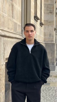 Svart fleecegenser med kvart-glidelås og teksturert overflate, brukt over en hvit t-skjorte. Mørke bukser kompletterer antrekket mot en steinvegg.