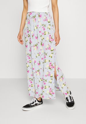Femme portant une longue jupe bleu clair à imprimé floral rose avec une fente latérale, associée à des baskets noires et blanches.