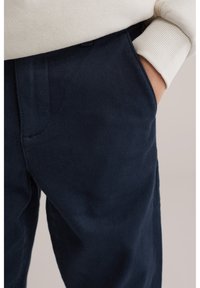 Oseba, oblečena v temno modre hlače, z roko v žepu in vidnim kremno belim rebrastim rokavom puloverja.