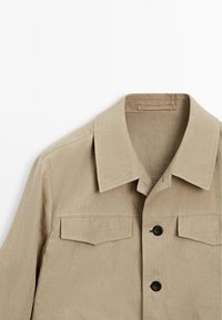 Beige Hemdblusenjacke mit einem Umlegekragen, zwei Vorderaschen und schwarzen Knöpfen. Hergestellt aus einem leichten Stoff mit glatter Textur.