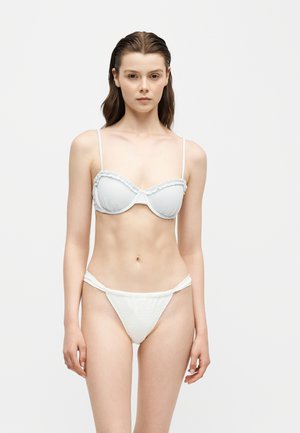 Ung kvinde med vådt hår iført en hvid todelt bikini med flæset top, står mod en ensfarvet lys baggrund.