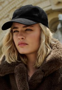 Cappello nero con visiera curvata, indossato sopra un colletto di pelliccia marrone. Il tessuto sembra testurizzato, in complemento ai capelli biondi mossi di media lunghezza.