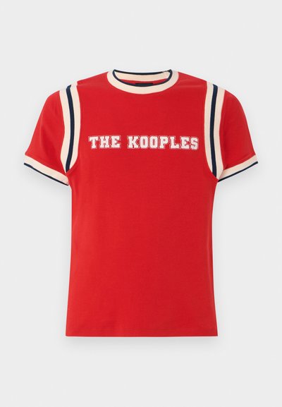 Punainen puuvillapaita, jossa on ribattu pyöreä pääntie, valkoiset ja tummansiniset raidat hartioilla, ja "THE KOOPLES" tulostettuna paksuilla valkoisilla kirjaimilla.