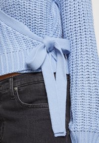 Pull en tricot bleu clair avec un motif texturé et un détail de nœud sur le côté, porté avec un jean en denim noir.