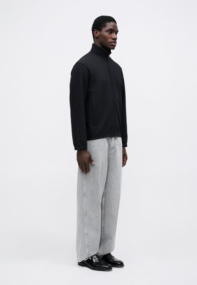 Čierna zapínacia bunda s vysokým golierom, skombinovaná s svetlošedými širokými džínsami a čiernymi topánkami. Minimalistický dizajn, hladká textúra.