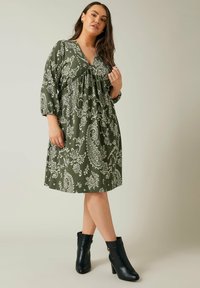 Vestido verde con estampado de cachemira, con escote en V y cintura fruncida, que presenta mangas de tres cuartos. Combinado con botas negras hasta el tobillo.