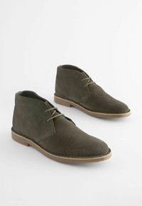 Botas de tobillo de ante verde con diseño de cordones, punta redonda y suela de goma marrón. Presenta una textura suave y un detalle de costura mínimo.