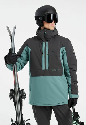 Herreskijakke i petrolblå og sort, med høj krave, frontlomme, justerbare manchetter og lynlåse. Modellen holder ski og bærer en hjelm.