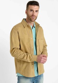 Veste boutonnée beige en tissu durable, présentant une poche poitrine et un col, portée sur une chemise bleu clair.