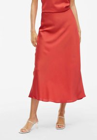 Falda midi de satén rojo con una textura suave y un ligero brillo. Forma en A con un dobladillo fluido. Combinada con tacones de tiras transparentes.