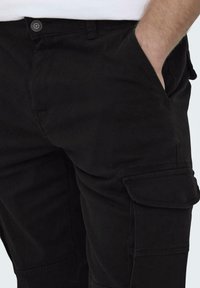 Pantalons cargo noirs avec une texture lisse, présentant des poches latérales, une fermeture à bouton et des accents cousus. Un design globalement fonctionnel.