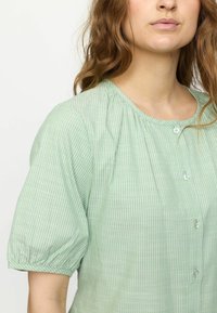 Ljust grön blus med vertikala vita ränder, korta puffärmar, rundad halsringning och knappdetaljer. Mjuk, strukturerad tyg.