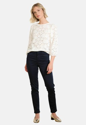 White lace long-sleeve top with a round neckline, paired with dark navy fitted pants and golden block-heel shoes.