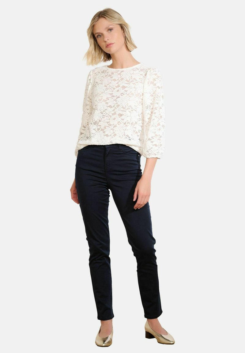 White lace long-sleeve top with a round neckline, paired with dark navy fitted pants and golden block-heel shoes.