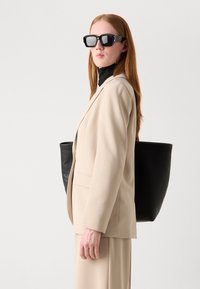 Beige tailored blazer over a black turtleneck, paired with a black tote bag. Model wears oversized black sunglasses and has long hair.