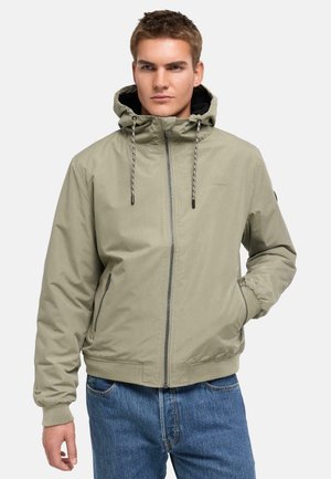 Ung mand iført en lysebeige hættejakke med lynlås og snore, kombineret med blå jeans, stående med den ene hånd i lommen.