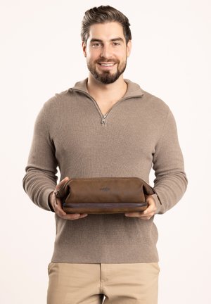 Brown leather toiletry bag with a zipper, displayed in hands. Ribbed beige sweater and neutral background accentuate the product.