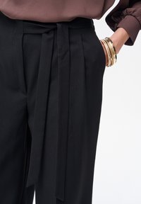 Czarne, dopasowane spodnie z szerokim pasem, ozdobione wiązanym paskiem. Gładka tkanina i wyraźne zakładki tworzą zdefiniowany wygląd. Złote bransoletki podkreślają całość.
