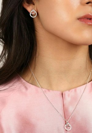Silverörhängen med cirkulär design, med en klar stendetalj. Matchande silverhalsband med sammanflätad cirkulär pendant. Rosa tygblus.
