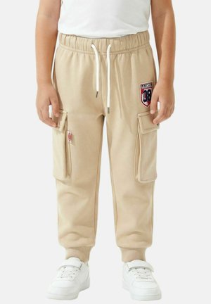 Enfant portant un jogger cargo beige avec détail en patch, cordon blanc et baskets blanches, debout devant un fond uni.