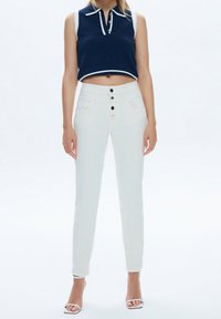 Jeans blancos de cintura alta con cinco botones, que presentan bolsillos delanteros y un ajuste slim. Combinados con un polo sin mangas de color marino con acentos en blanco.