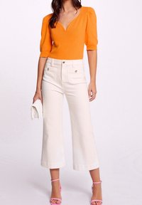 Haut en tricot orange avec un col en V et des manches bouffantes, associé à un pantalon large crème taille haute. Les accessoires comprennent une pochette blanche et des talons roses.