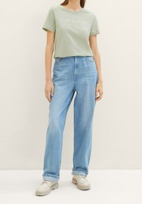 Lys grøn t-shirt med "TOM TAILOR" trykt, parret med løstsiddende, lyseblå denim jeans og beige chunky sneakers.