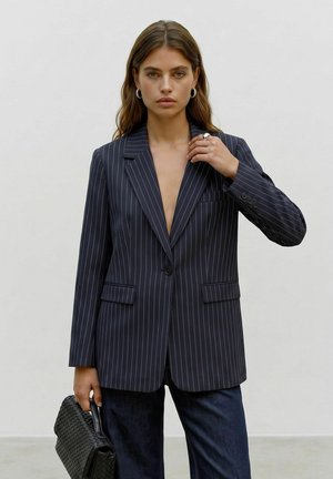 Femme aux longs cheveux portant un blazer sombre à fines rayures et un pantalon foncé, tenant un sac à main tissé noir sur un fond gris uni.