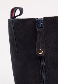 Parte superior de bota de ante negro con un zipper azul marino y un acento de botón dorado. Cuenta con un tirador en bucle en la parte superior. Textura suave.