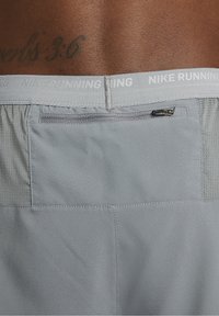Gros plan du bas du dos d'une personne portant un short Nike Running gris clair avec une poche zippée et un tatouage visible au-dessus de la ceinture.