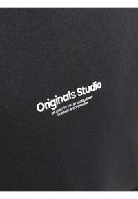 Svart tyg med en slät yta har texten "Originals Studio" i vitt. Inkluderar detaljer som anger varumärket och designens ursprung.