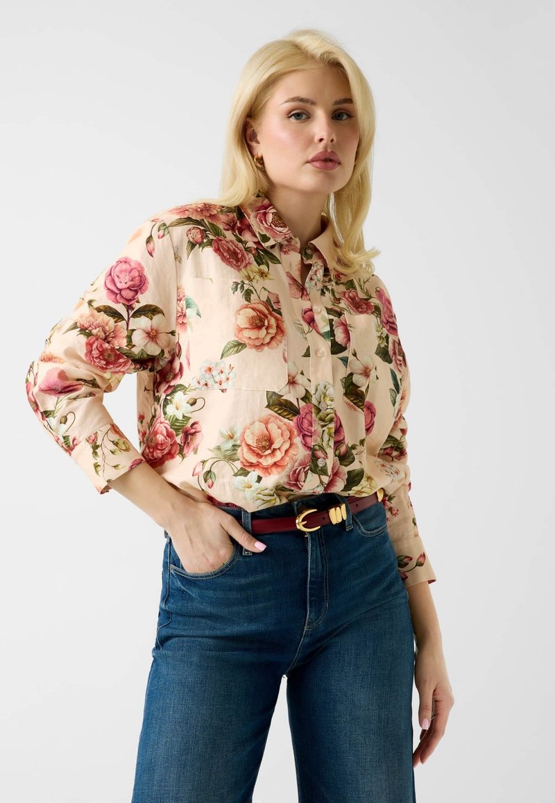 Femme blonde portant une chemise claire à fleurs boutonnée, rentrée dans un jean bleu avec une ceinture rouge, debout une main dans la poche sur fond blanc.