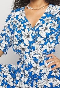 Květinové šaty v modré barvě s bílým a černým květinovým potiskem, krátké rukávy, výstřih do V a zavázaný pas. Hladký textilní povrch.