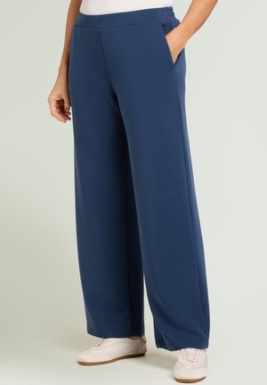 Person trägt weit geschnittene marineblaue Hose mit elastischem Bund und Seitentaschen, kombiniert mit weißen Turnschuhen.