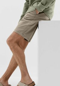 Shorts bej dintr-un material ușor, cu croială dreaptă. Asortați cu sandale de culoare deschisă. Fundalul este simplu și minimalist.