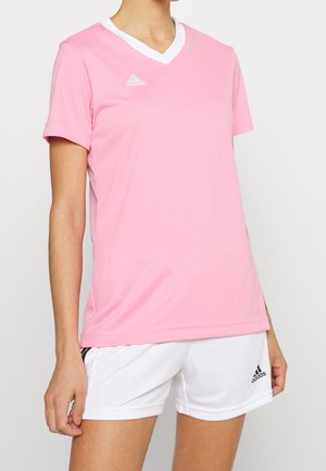 Persoon draagt een roze Adidas V-hals T-shirt met korte mouwen en witte Adidas shorts, staand met de arm ontspannen naast het lichaam.