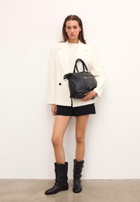 Fekete bőr kézitáska, strukturált forma, texturált felület, dupla felső fogantyúkkal és ezüst színű fémrészekkel, amit egy modell tart egy világos blézerben és sortban.